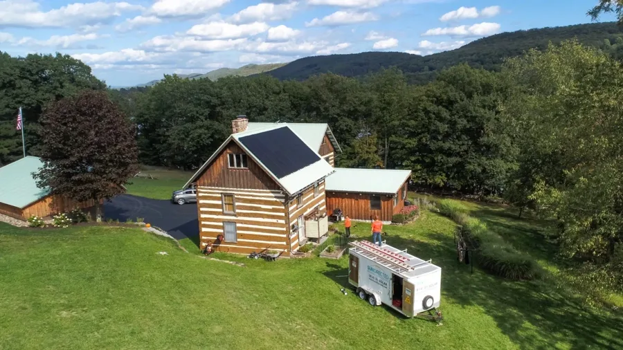 Aerial of a log cabin with rooftop solar and a Sun Directed work trailer on the lawn.